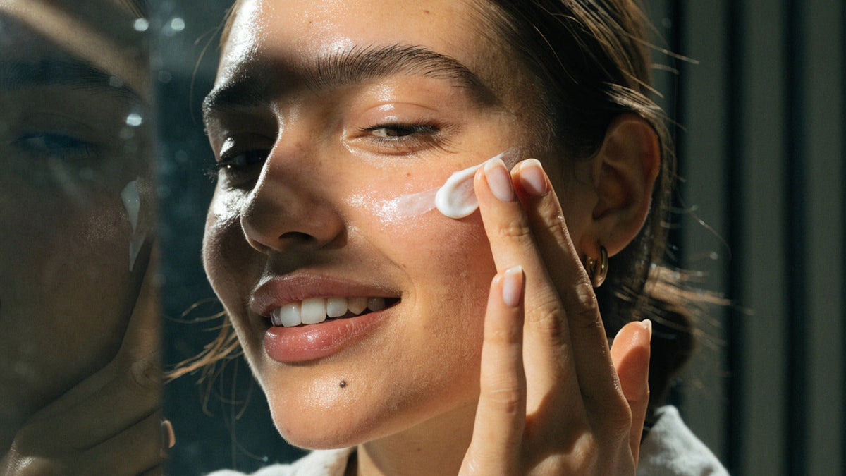 Person applying cream to the cheek beside a mirror in natural light.